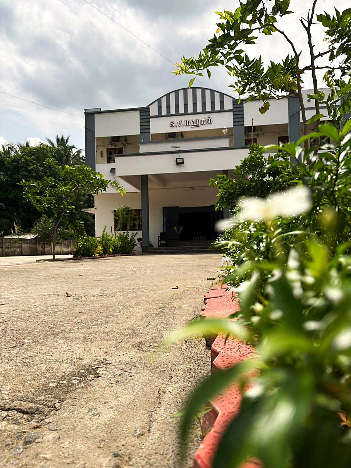 A two-storey white building with grey trim, its name board above the portico, seen across the forecourt through flowering shrubs.