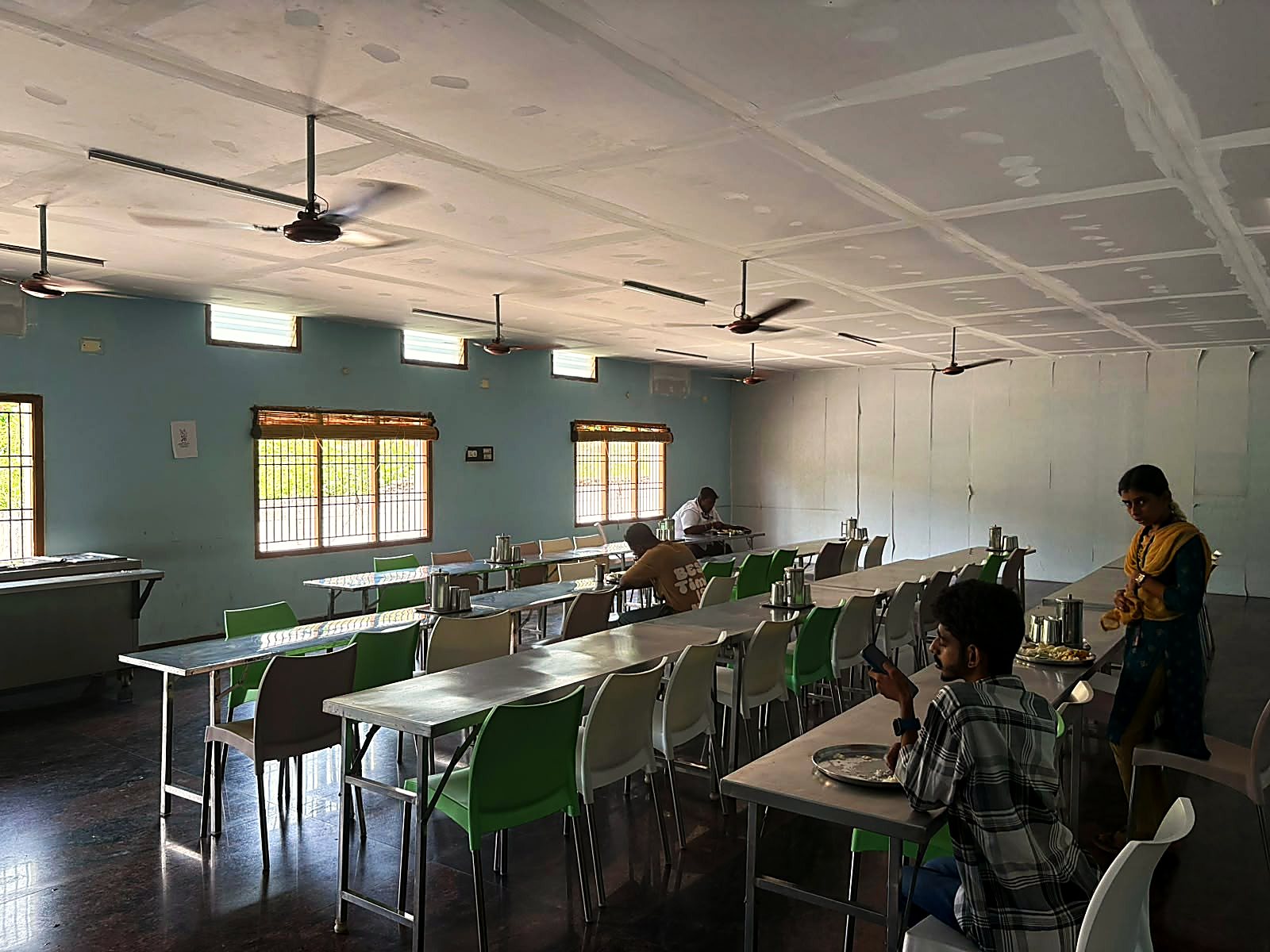 The canteen, with long steel tables, green and white chairs, and a few people eating lunch.