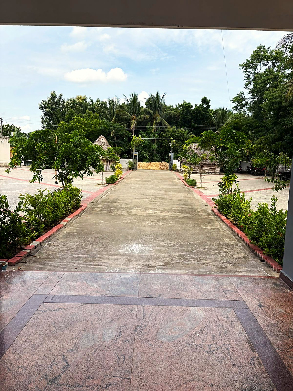 The view out from the office entrance: a concrete drive with a kolam chalked on it, flanked by planted beds and thatched huts.