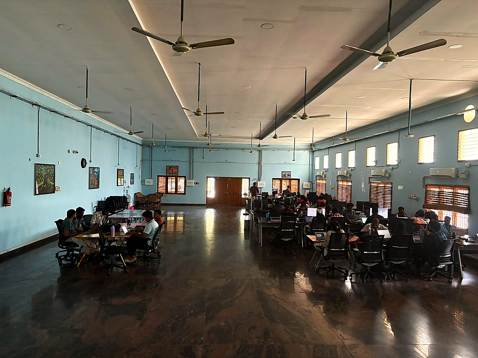 The main hall at the Kurichi office: a high, pale-blue room with ceiling fans, two long rows of desks, and people working at laptops.